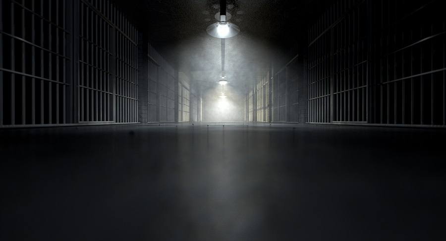 A concept image of an eerie corridor in a prison at night showing jail cells dimly illuminated by various ominous lights