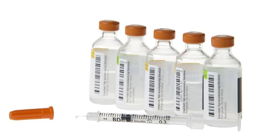 Medication bottles vials with injectable medication lined up in a row with a 40 unit per milliliter syringe lying in front with one drop of medication coming out of the tip of the needle. Cap on side
